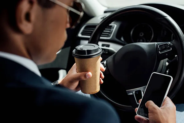 Back view of african american businessman in suit and sunglasses using smartphone and drinking coffee in car — Stock Photo