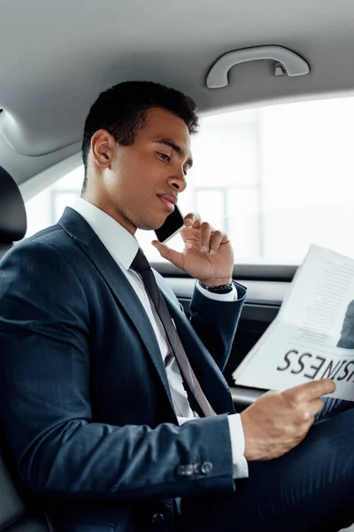 African american businessman reading business newspaper and talking on smartphone in car — Stock Photo