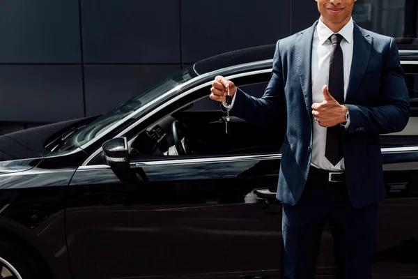 Ausgeschnittene Ansicht eines afrikanisch-amerikanischen Mannes im Anzug, der Autoschlüssel in der Hand hält und den Daumen in der Nähe eines schwarzen Autos zeigt — Stockfoto