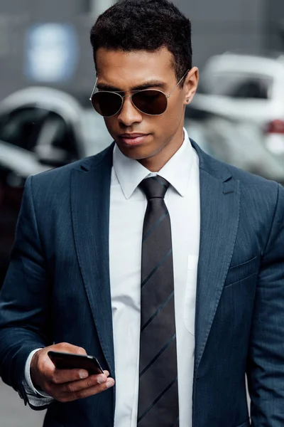 Guapo hombre de negocios afroamericano en traje y gafas de sol usando smartphone - foto de stock