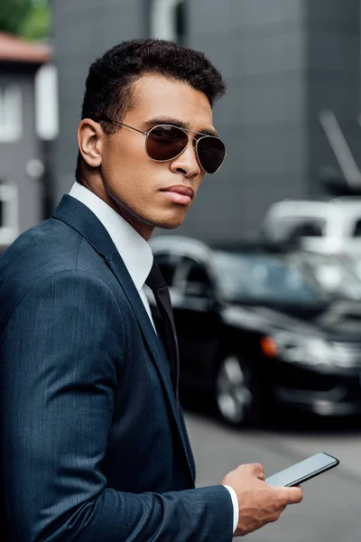 Beau et confiant homme d'affaires afro-américain en costume et lunettes de soleil en utilisant smartphone — Photo de stock