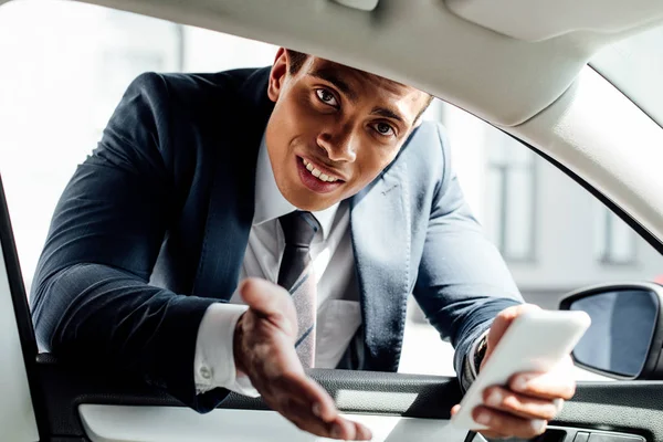 Homme d'affaires afro-américain en costume regardant dans la voiture tout en tenant smartphone — Photo de stock