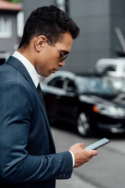 Vista lateral del hombre de negocios afroamericano guapo y confiado en traje y gafas de sol usando teléfono inteligente - foto de stock