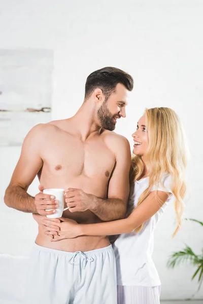 Schöne Frau umarmt Mann mit Kaffeetasse zu Hause in der Früh — Stockfoto