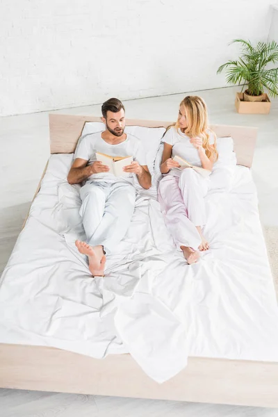 Vue grand angle du couple en pyjama se détendre au lit avec des livres à la maison — Photo de stock