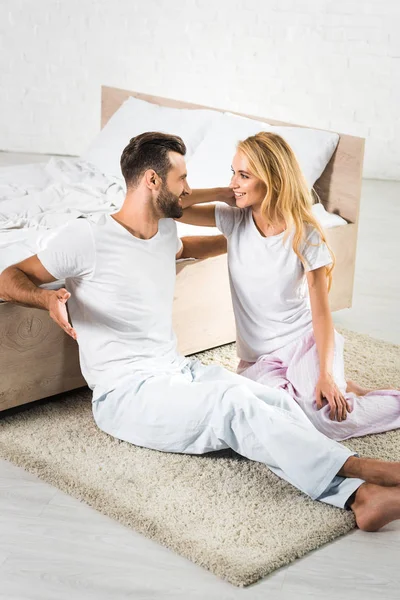 Hermosa pareja sentada en la alfombra y hablando cerca de la cama en casa - foto de stock