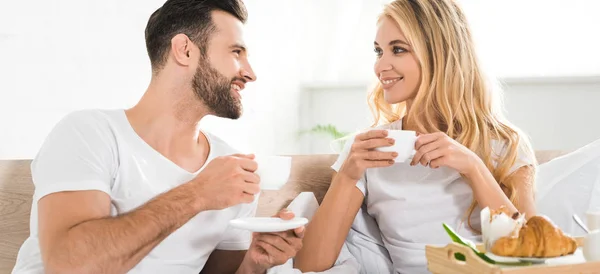 Plano panorámico de hermosa pareja con tazas durante el desayuno en la cama por la mañana - foto de stock