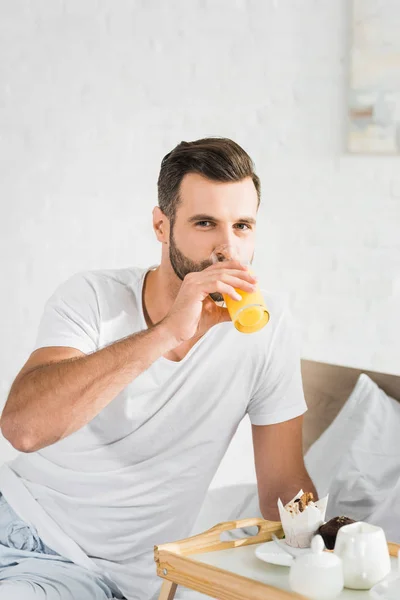 Hombre guapo bebiendo jugo de naranja cerca de bandeja de comida durante el desayuno en casa - foto de stock