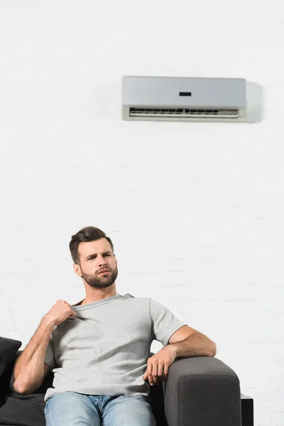 Handsome man sitting on couch and suffering from heat under air conditioner at home — Stock Photo