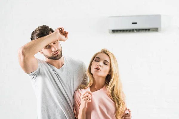 Pareja limpiando la frente y sufriendo de calor bajo el aire acondicionado en casa - foto de stock