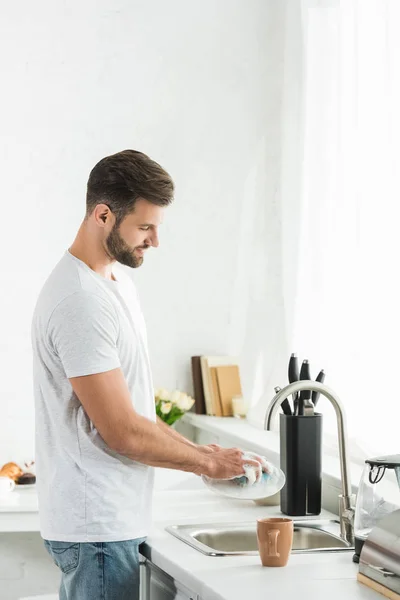 Hombre guapo lavando platos en la cocina por la mañana - foto de stock