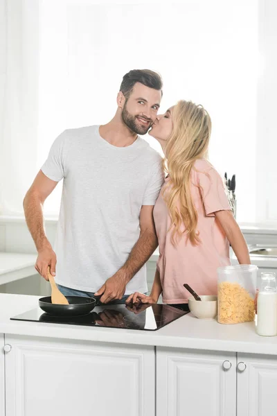 Жінка цілує чоловіка, готуючи сніданок на кухні — стокове фото