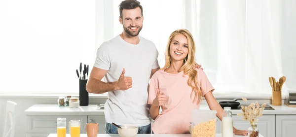 Plano panorámico de hermosa pareja mostrando los pulgares hacia arriba durante el desayuno en la cocina - foto de stock