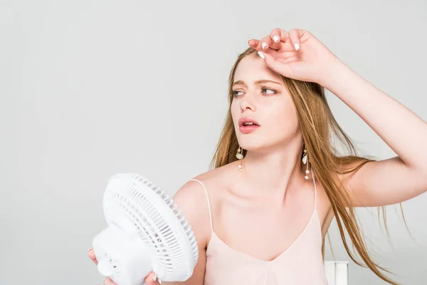 Pretty young woman suffering from heat and holding Electric Fan isolated on grey — Stock Photo
