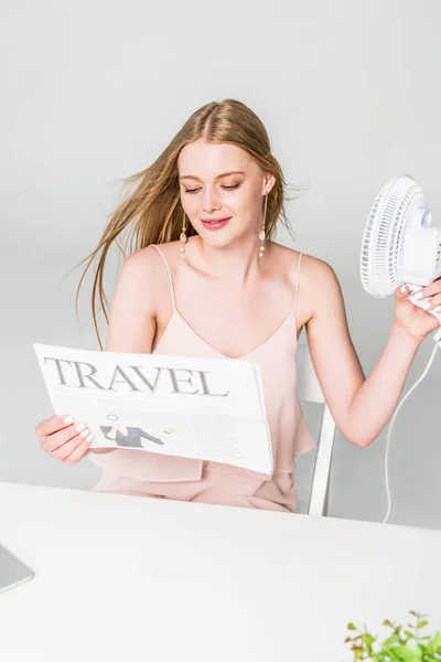 Красивая молодая женщина с электрическим вентилятором и газетой путешествия страдает от жары на сером — стоковое фото