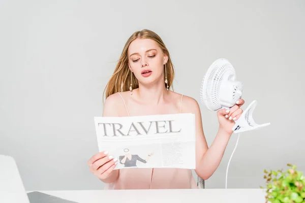 Красивая молодая женщина с электрическим вентилятором и туристической газеты страдают от жары изолированы на сером — стоковое фото