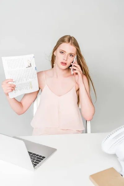 Beautiful girl suffering from heat, waving newspaper and talking on smartphone on grey — Stock Photo