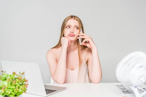 Hermosa chica hablando en el teléfono inteligente y sufriendo de calor en el escritorio de la computadora aislado en gris - foto de stock