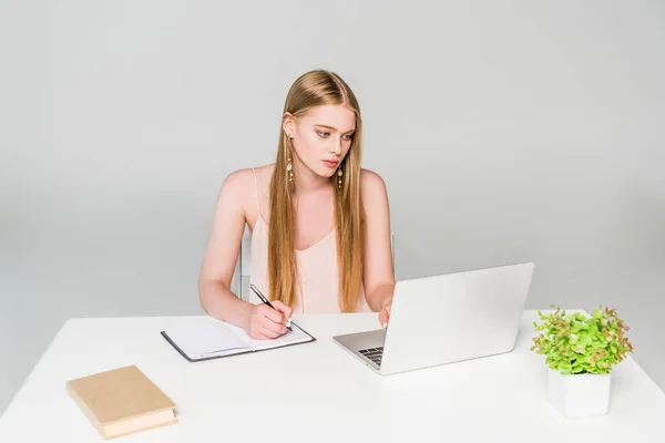 Beautiful girl sitting at computer desk and writing in notebook on grey — Stock Photo