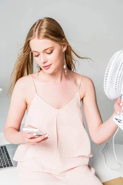 Красива молода жінка з електричним вентилятором страждає від спеки і використовує смартфон на сірому — стокове фото