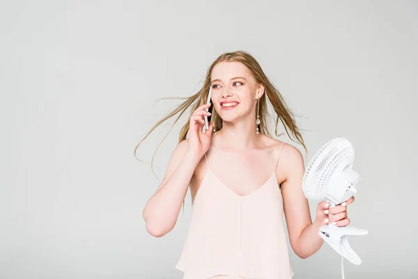 Красива молода жінка з електричним вентилятором страждає від спеки і розмовляє на смартфоні ізольовано на сірому — стокове фото