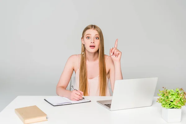Красива дівчина сидить за комп'ютерним столом і показує жест ідеї ізольовано на сірому — стокове фото