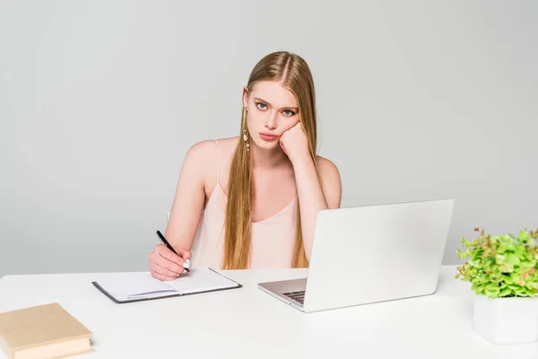Chica disgustado sentado en el escritorio de la computadora y escribir en cuaderno en gris - foto de stock