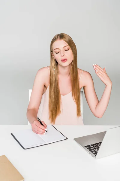 Beautiful girl sitting at computer desk, writing in notebook and suffering from heat on grey — Stock Photo