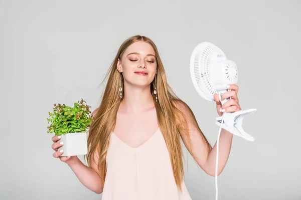 Bela jovem com ventilador elétrico e vaso de plantas que sofrem de calor isolado em cinza — Fotografia de Stock
