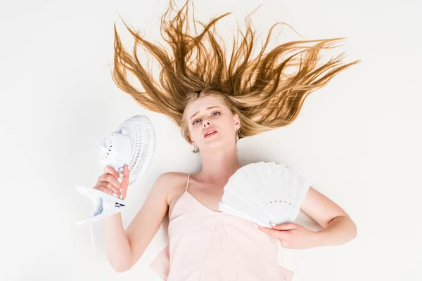 Вид зверху красива молода жінка з електричним вентилятором і ручним вентилятором, що страждає від спеки на білому — стокове фото