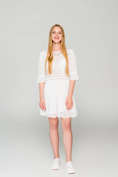 Красива усміхнена дівчина в білій сукні дивиться на камеру на сірому — стокове фото