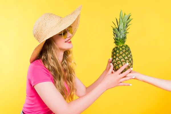 Hermosa chica en sombrero de paja dando piña a la mujer aislado en amarillo - foto de stock