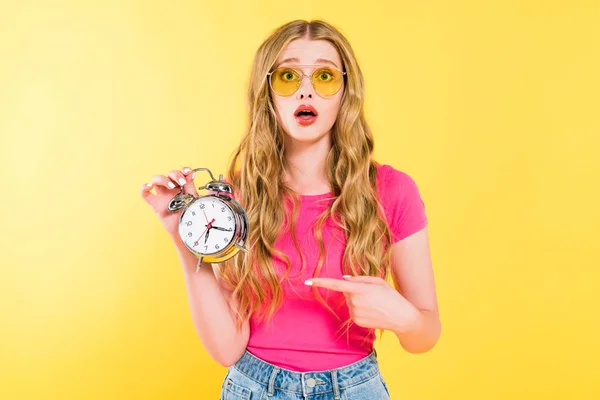 Beautiful surprised girl in sunglasses pointing with finger at alarm clock On yellow — Stock Photo