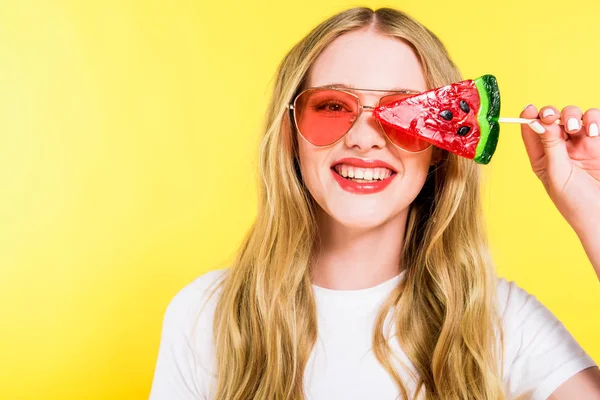 Hermosa chica feliz en gafas de sol con piruleta en forma de sandía aislado en amarillo - foto de stock