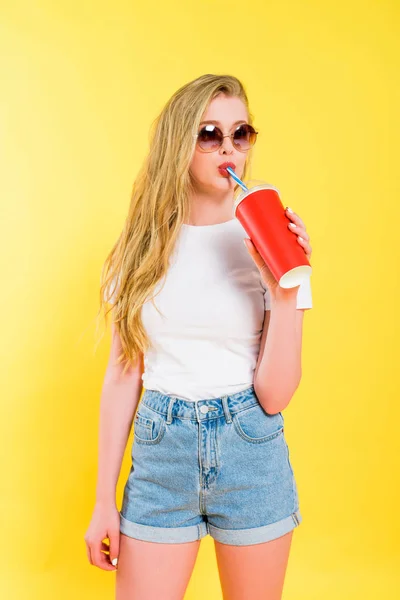 Belle fille dans des lunettes de soleil boire soda de tasse jetable Sur jaune — Photo de stock