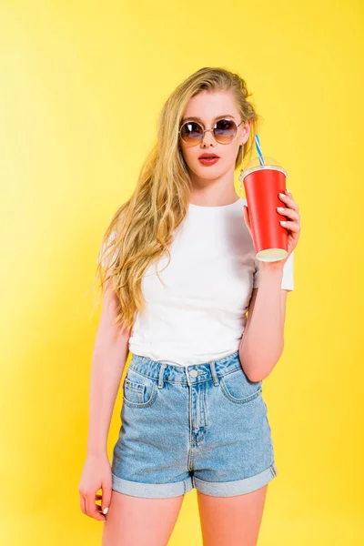 Belle fille dans des lunettes de soleil avec boisson dans une tasse jetable Sur jaune — Photo de stock