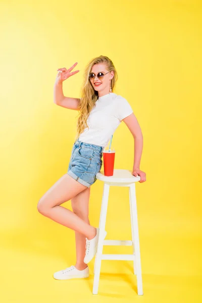 Schönes Mädchen mit Sonnenbrille in der Nähe Stuhl mit Limonade zeigt Friedenszeichen auf gelb — Stockfoto