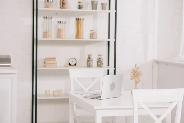 Cuisine légère avec ordinateur portable sur table blanche, chaises et rack avec bocaux — Photo de stock