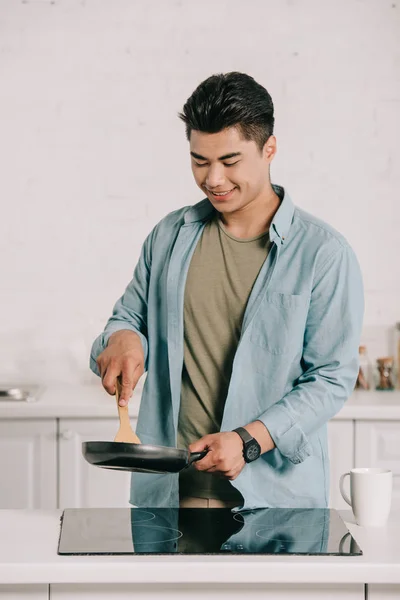 Smiling asian man cooking breakfast on frying pan in kitchen — Stock Photo