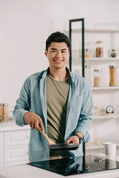 Fröhlicher asiatischer Mann lächelt in die Kamera, während er das Frühstück auf der Pfanne zubereitet — Stockfoto