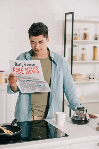 Bello asiatico uomo lettura falso notizie giornale mentre in piedi a cucina tavolo vicino piano di cottura, caffettiera e tazza — Foto stock