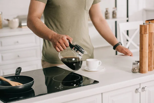 Частичный вид азиатского человека наливая кофе в чашку, стоя возле поверхности приготовления пищи на кухне — стоковое фото
