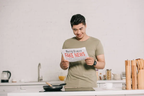 Smiling asian man reading fake news newspaper while standing near cooking surface in kitchen — Stock Photo