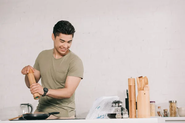 Fröhlicher asiatischer Mann kocht Frühstück, während er Zeitung in der Küche liest — Stockfoto