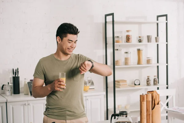 Alegre asiático homem olhando para assistir enquanto segurando vidro de suco de laranja — Fotografia de Stock