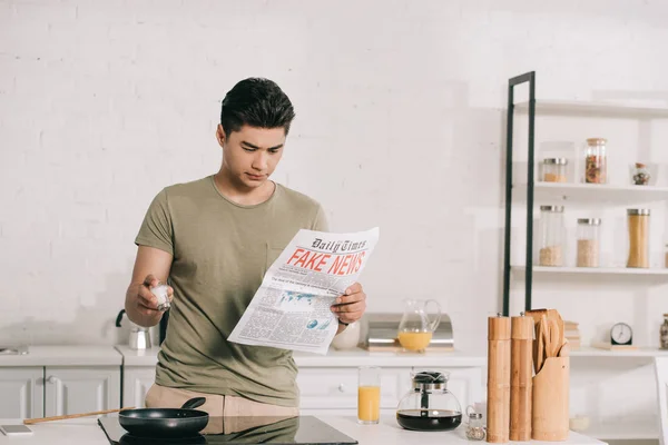 Handsome asian man preparing breakfast while reading fake news newspaper in kitchen — Stock Photo