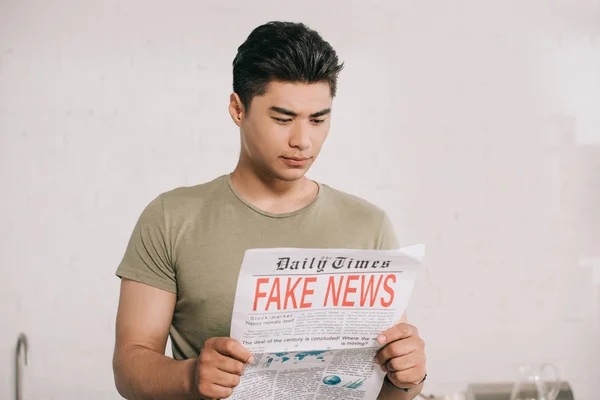 Jovem, atencioso asiático homem lendo falso notícias jornal na cozinha — Fotografia de Stock