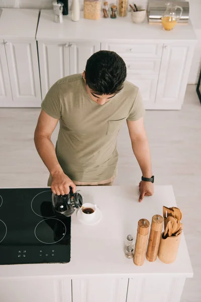 Vista aérea del joven que vierte café en taza mientras está de pie cerca de la superficie de cocción en la cocina - foto de stock