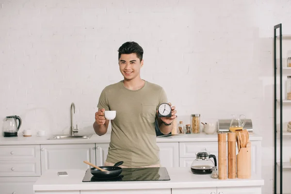 Cheerful asian man holding coffee cup and alarm clock while standing near cooking surface with frying pan — Stock Photo