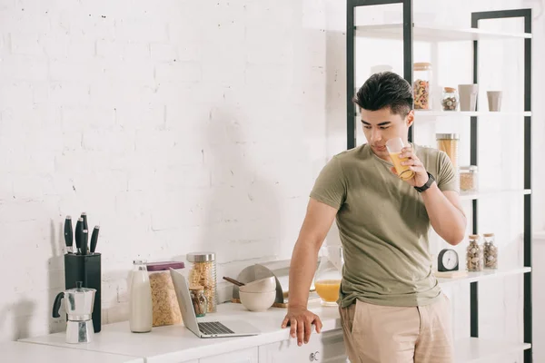 Schöner asiatischer Mann trinkt Orangensaft, während er Laptop auf Küchentisch betrachtet — Stockfoto
